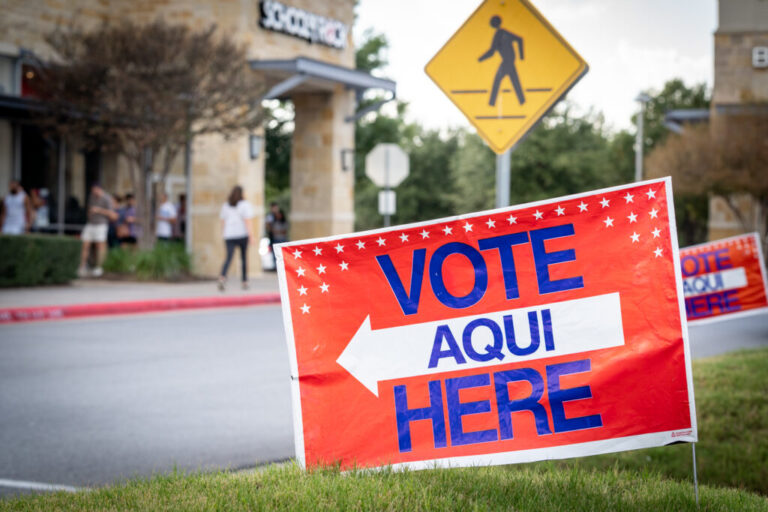 A voting sign is seen near a pedestrian walk way sign with building in the background.